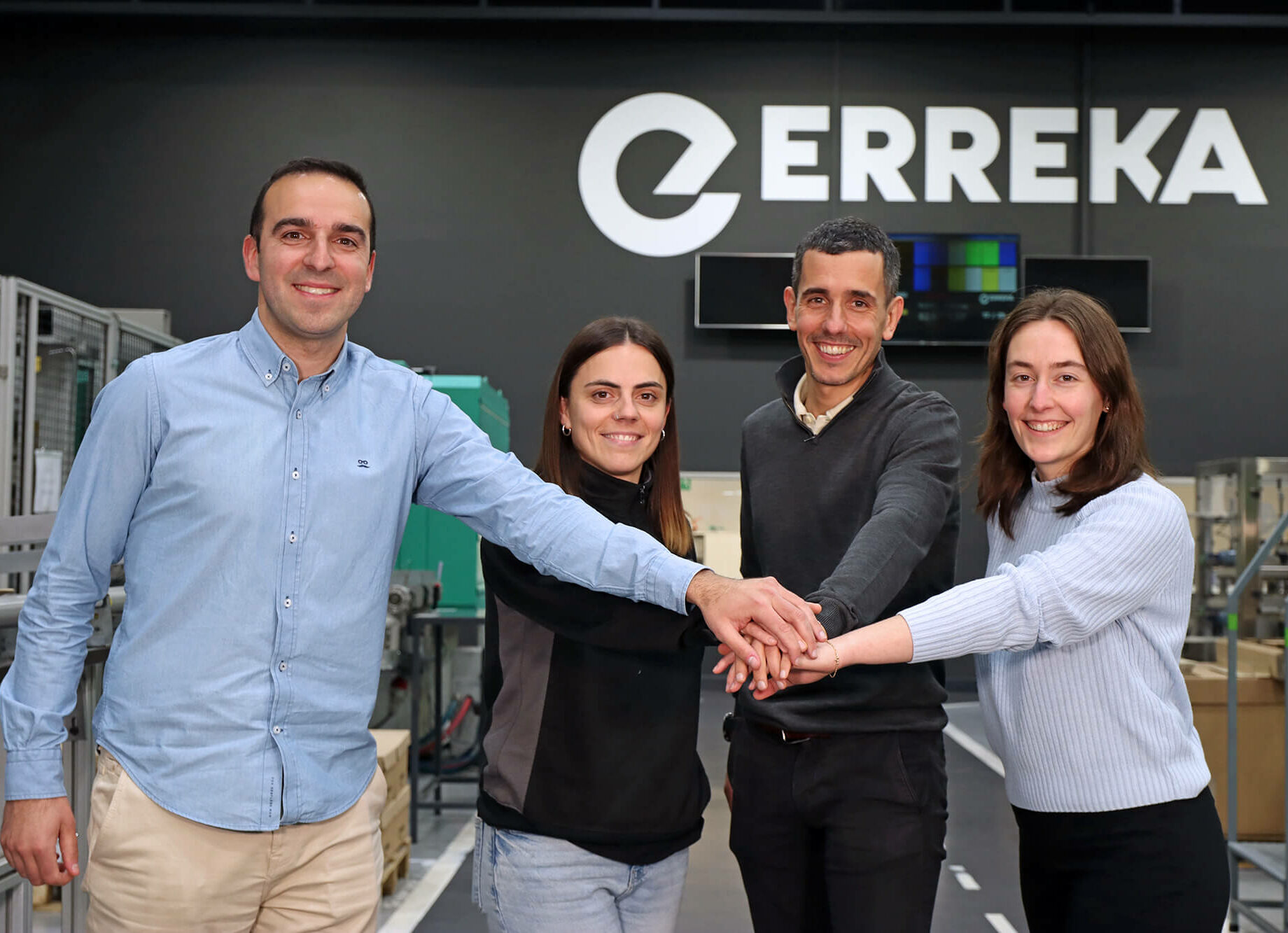 Four colleagues in a factory smile as they join hands in the center, with the ERREKA logo behind them.