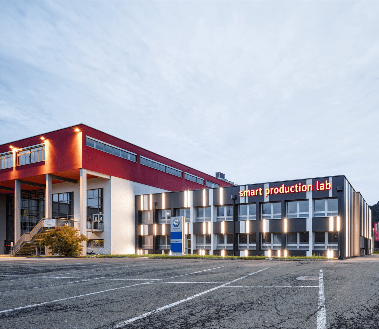 Modern office complex with a red upper level, glass windows, and a grey wing reading 'smart production lab'; empty parking lot in foreground.