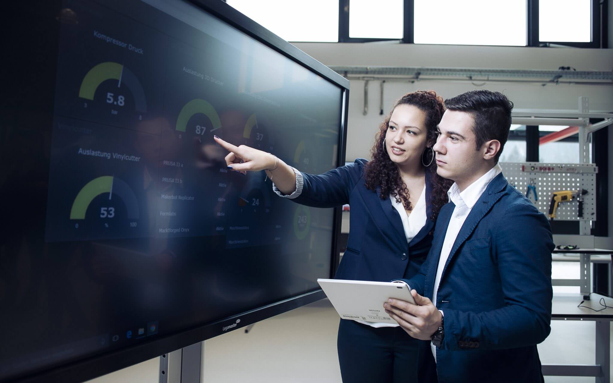 Two professionals in business attire point at a large touchscreen displaying gauges and charts.