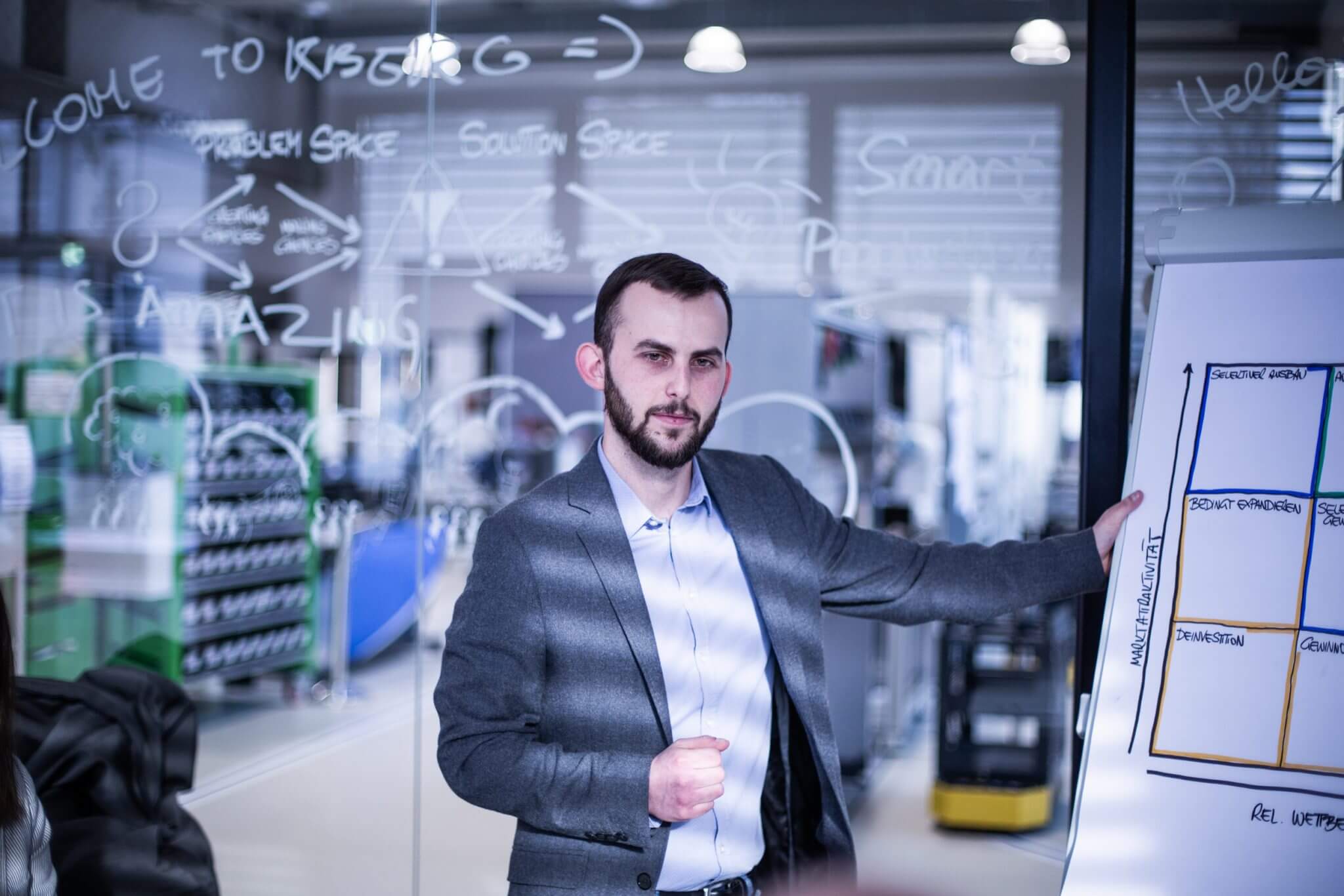 Man in a suit presenting a whiteboard with diagrams in a glass-walled meeting room.