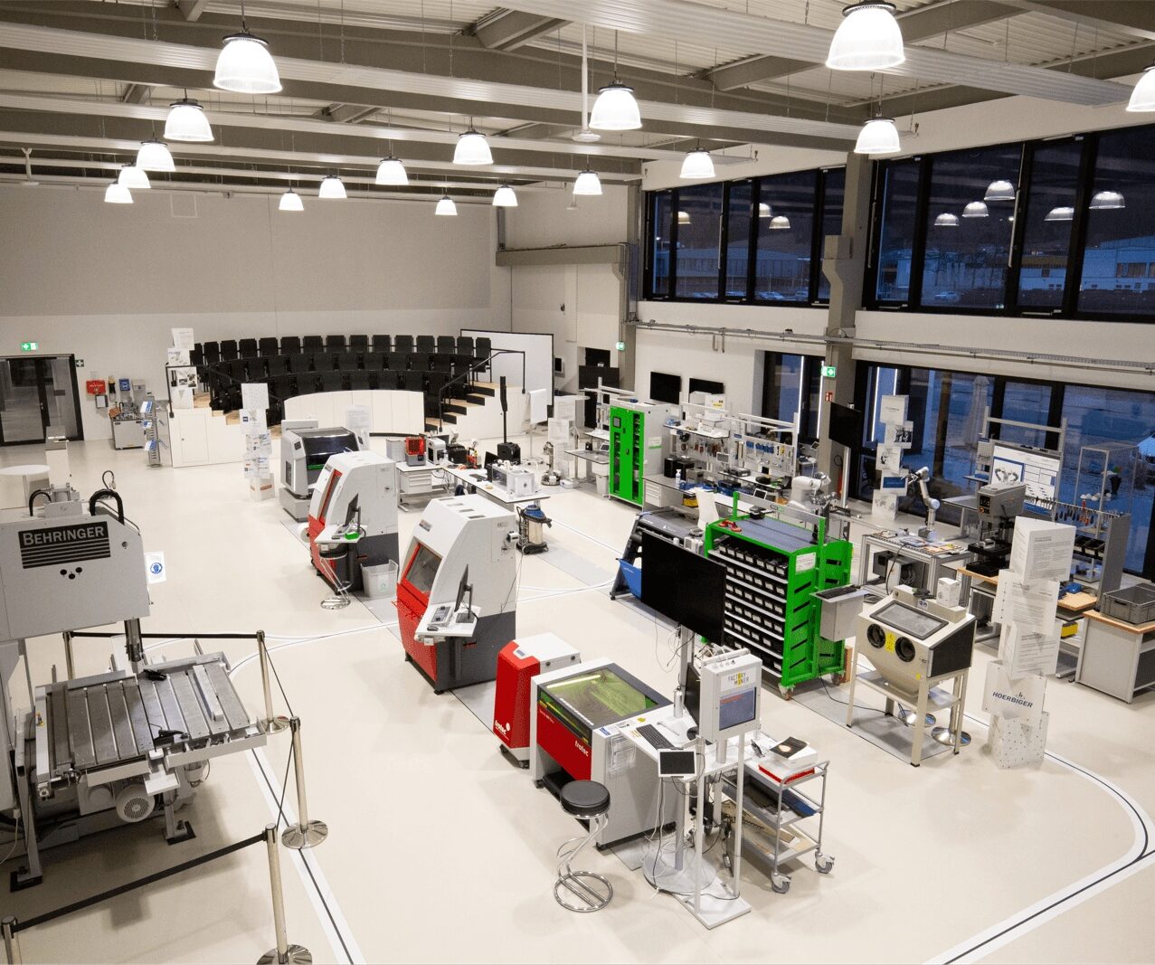Factory-style laboratory with multiple CNC/milling machines and workstations arranged around a white floor, bright ceiling lights, and a black-tier seating area in the back.