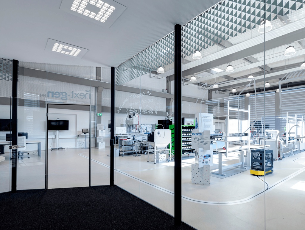 Open modern workshop behind glass walls, with workstations, shelving, and industrial equipment in a bright, organized space.—
