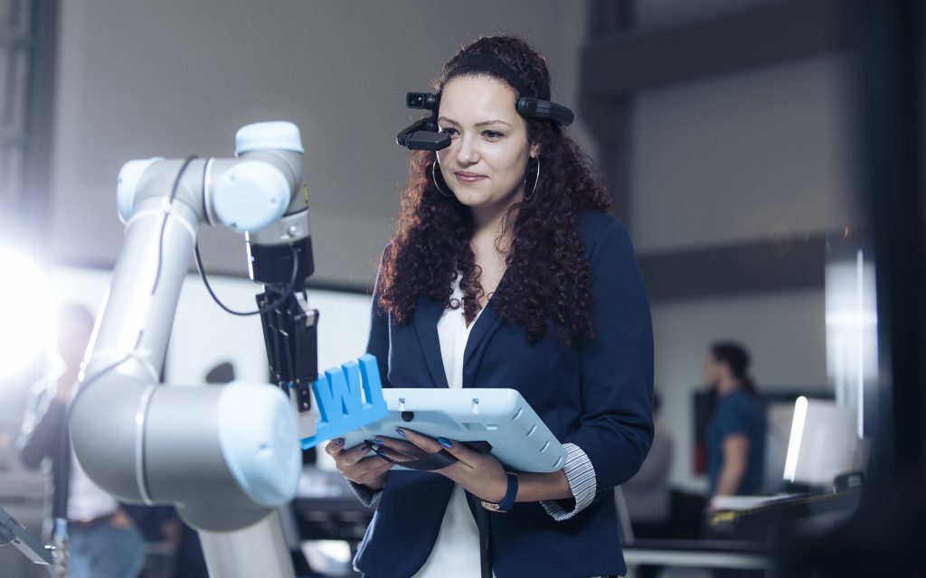 Woman in a navy blazer holds a tablet beside a robotic arm in a lab, wearing a head-mounted device.