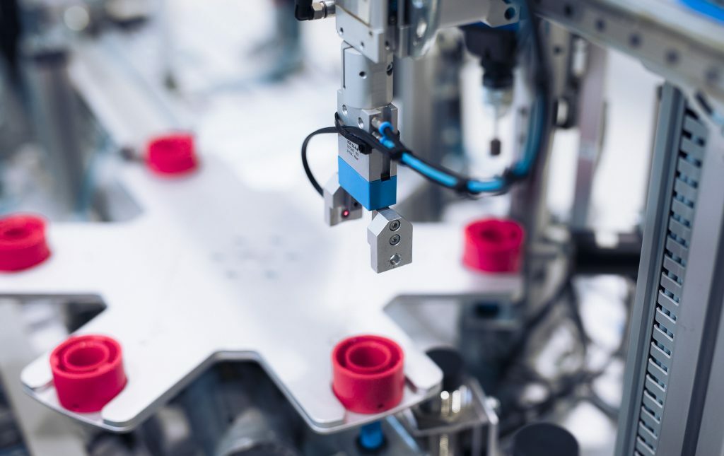 Close-up of automated manufacturing equipment with a robotic gripper above pink-red cups on a white conveyor belt, showing precision tooling.