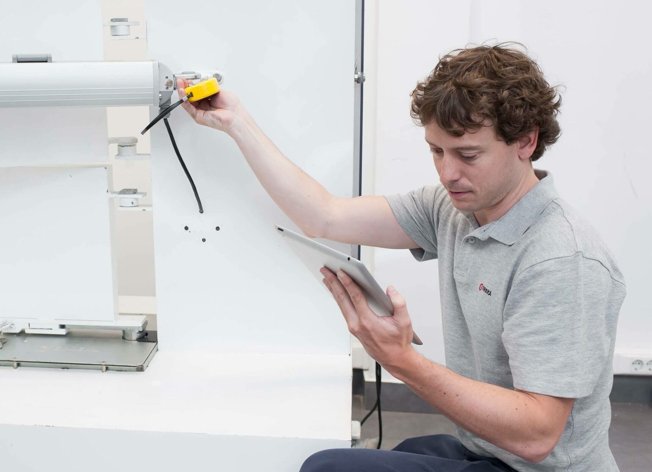 Man in a gray polo uses a yellow handheld device on a large white industrial machine, holding a tablet in the other hand.