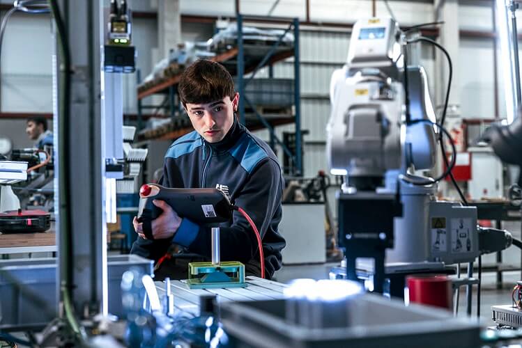 Young technician in a blue-gray jacket studies a tablet amid factory machinery and a robotic arm in the backgroundide.