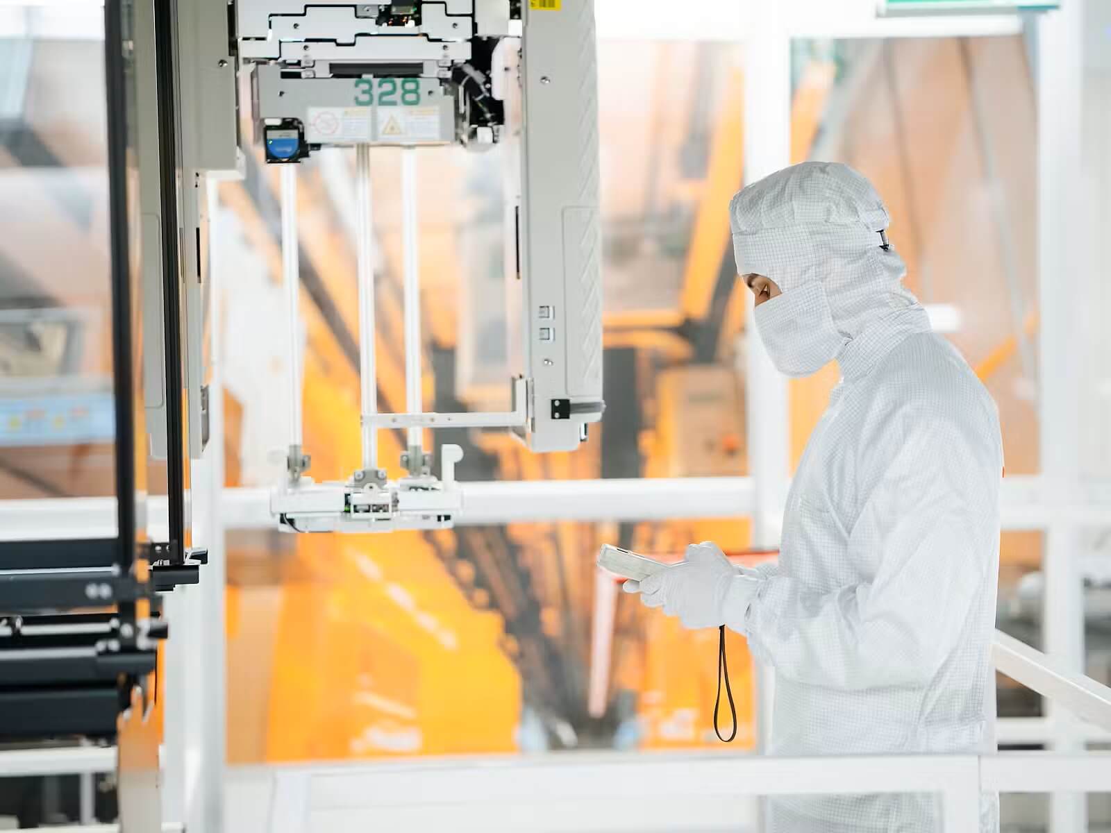 Worker in cleanroom suit operating a large automated manufacturing robot with a tablet nearby.