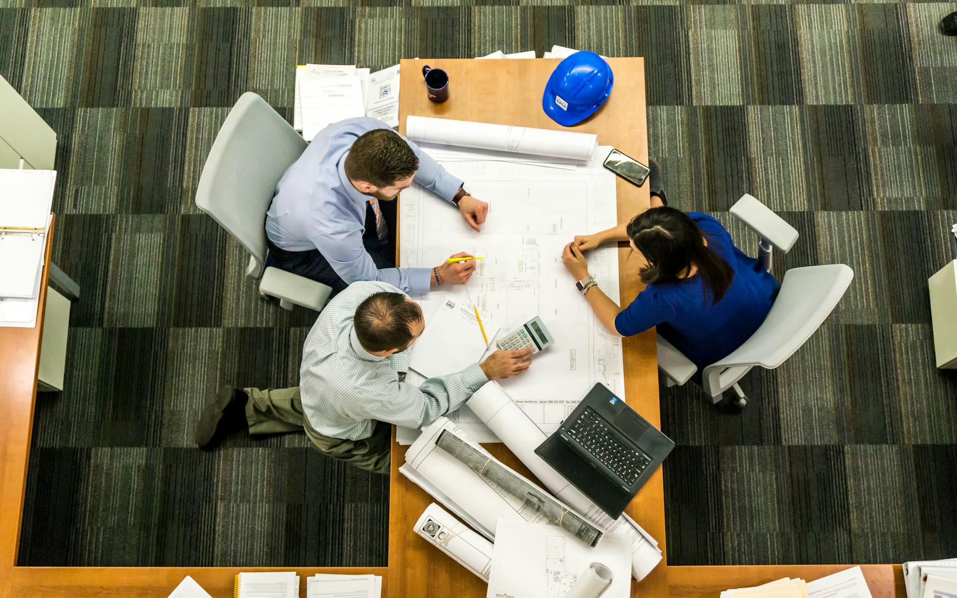 Overhead view of three professionals at a wooden table reviewing blueprints, with a blue hard hat, rolled plans, laptop, and phone nearby.