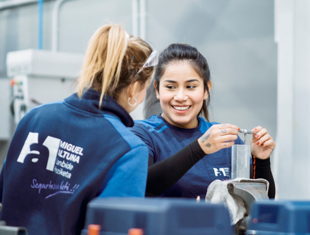 Two women in blue work uniforms collaborate at a workshop, one hands a small tool to the other near a metal vise panel.