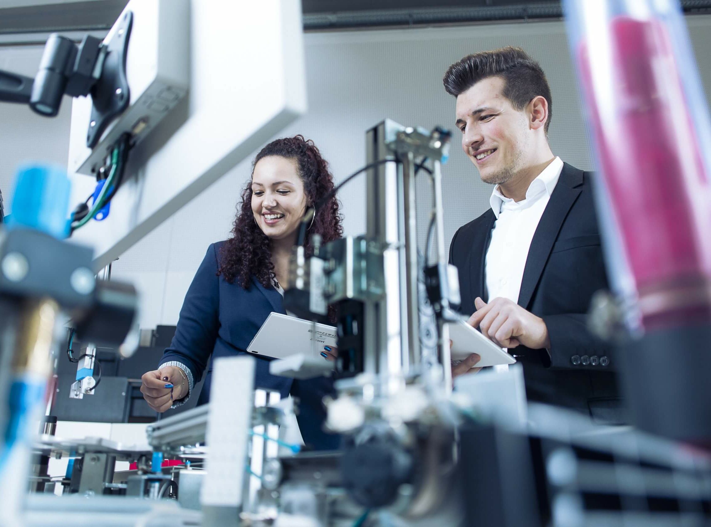Two professionals in business attire examine lab equipment, one holding a tablet and the other a clipboard, smiling as they work.