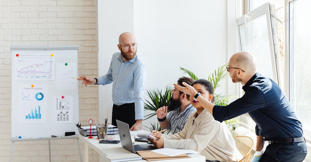 Bald man in a blue shirt presents charts on a whiteboard as colleagues discuss in a bright office.
