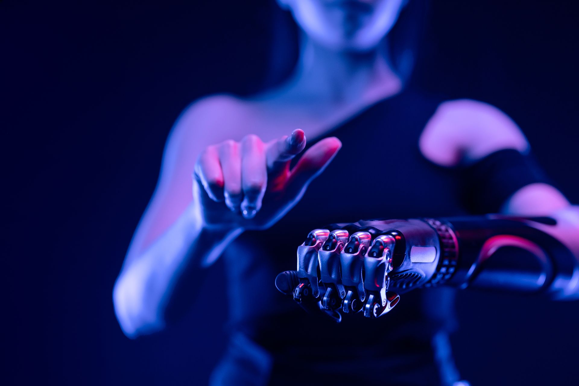 Close-up of a metallic prosthetic hand reaching toward a pointing finger, under blue-purple lighting.