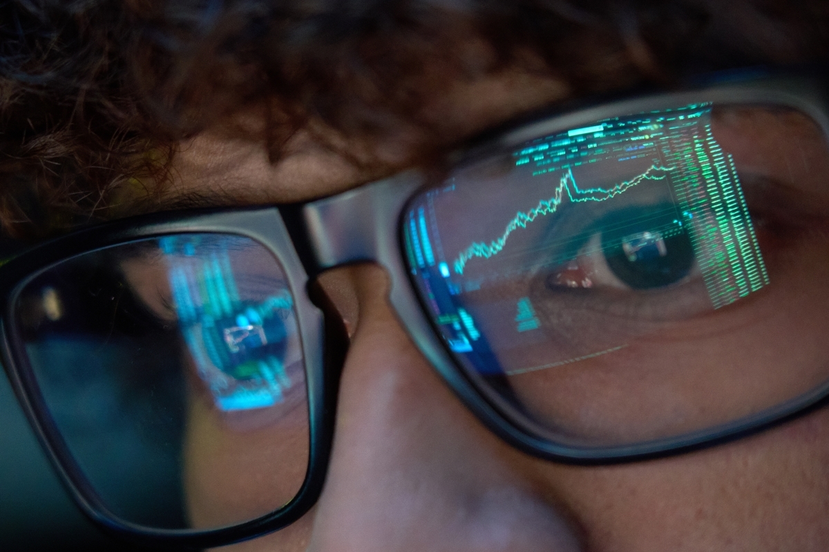 Close-up of a person wearing glasses, with reflections of stock market charts and green code on the lenses.