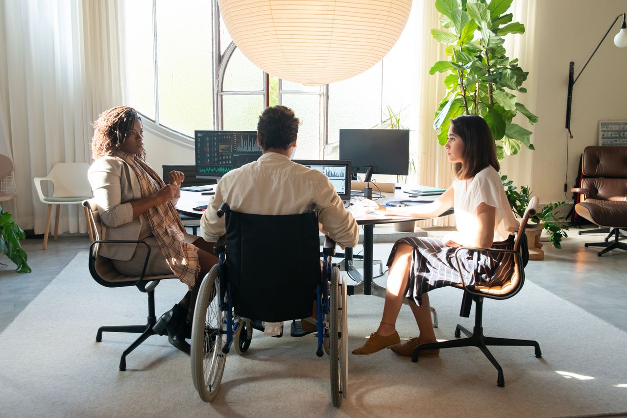 Team meeting in an airy office with three people around a desk; one person in a wheelchair facing monitors displaying charts and graphs.