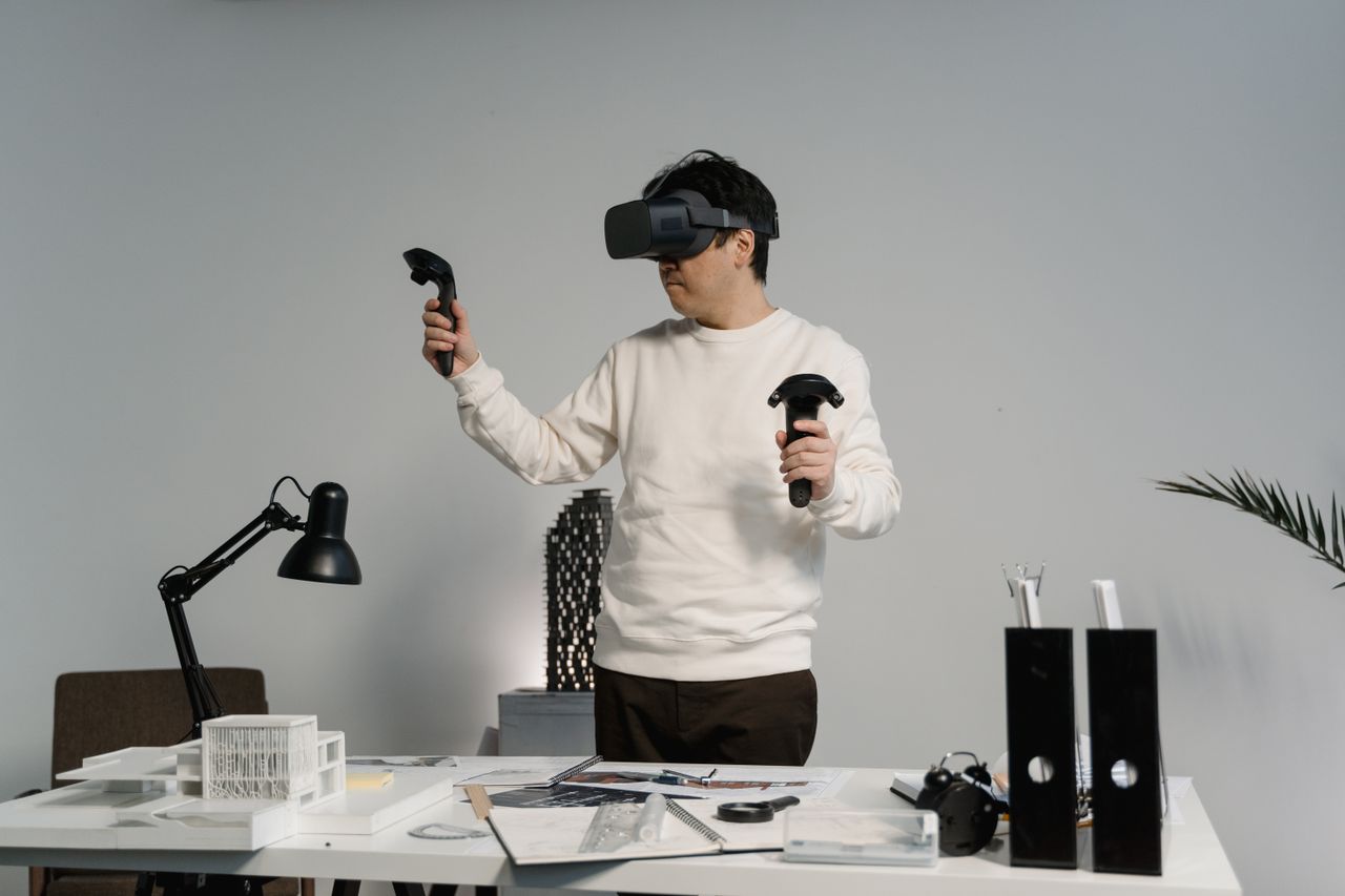 Person wearing a VR headset and holding two controllers at a cluttered desk in a bright workspace.